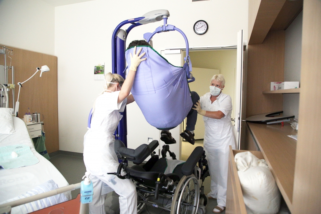 Pflegerinnen setzen Patient mit Hilfe eines Gerätes in einen Rollstuhl